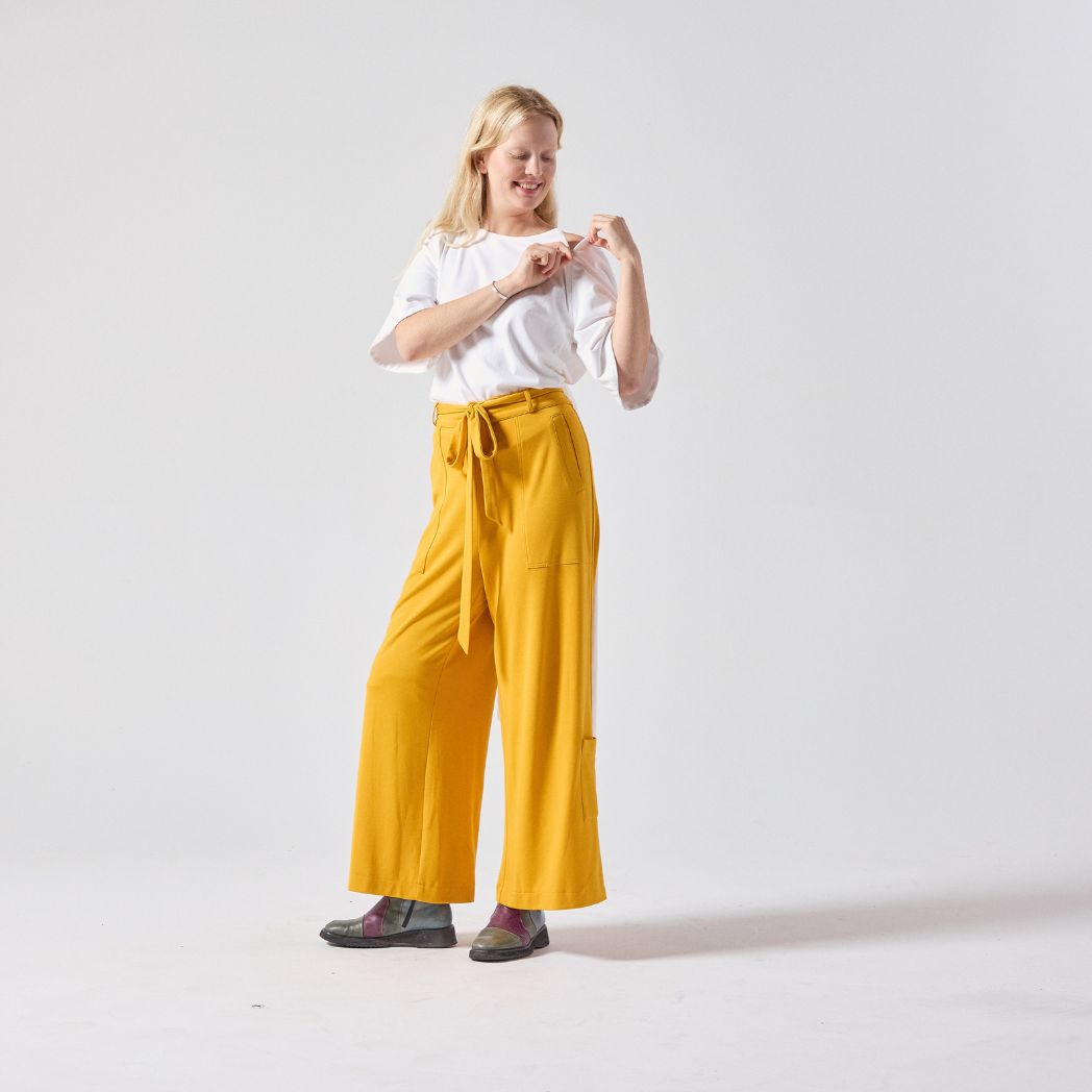 A white woman with blonde hair poses standing, looking away from the camera. She is wearing a white Jersey wrap top paired with yellow culottes and her own boots. She is standing with her hands unzipping the right front zipper of the wrap top. She is standing against a light grey background