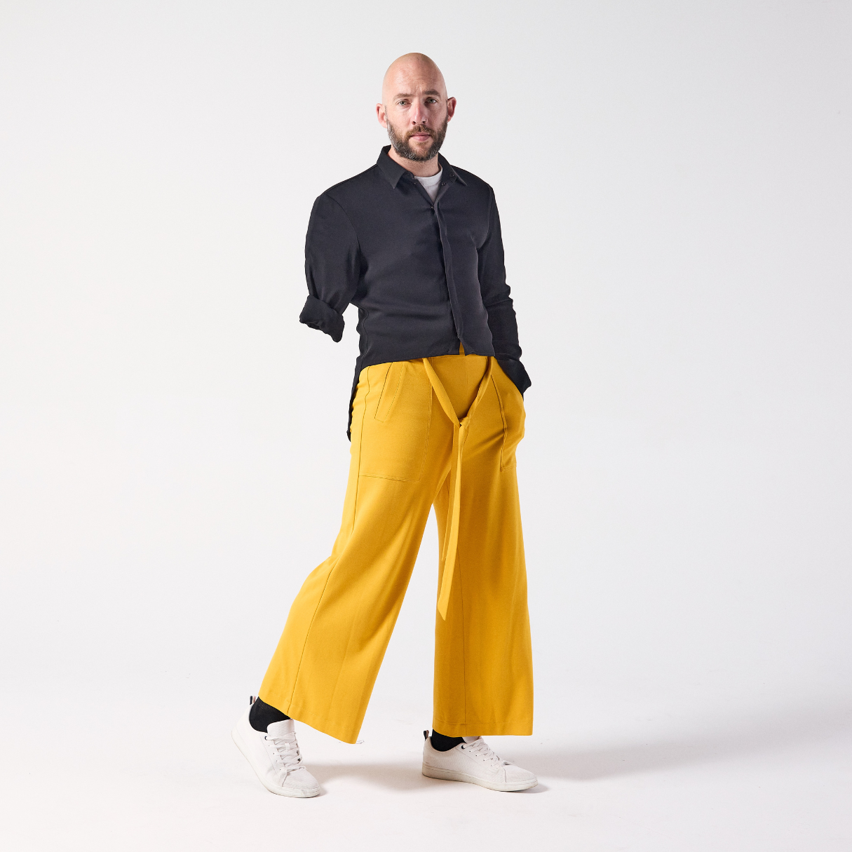 A white man is posing, looking at the camera. He is wearing jersey culottes in mustard yellow paired with a black shirt and white sneakers. He is standing with one hand in his pocket and his leg posed slightly outwards.