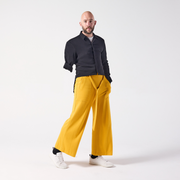 A white man is posing, looking at the camera. He is wearing jersey culottes in mustard yellow paired with a black shirt and white sneakers. He is standing with one hand in his pocket and his leg posed slightly outwards.