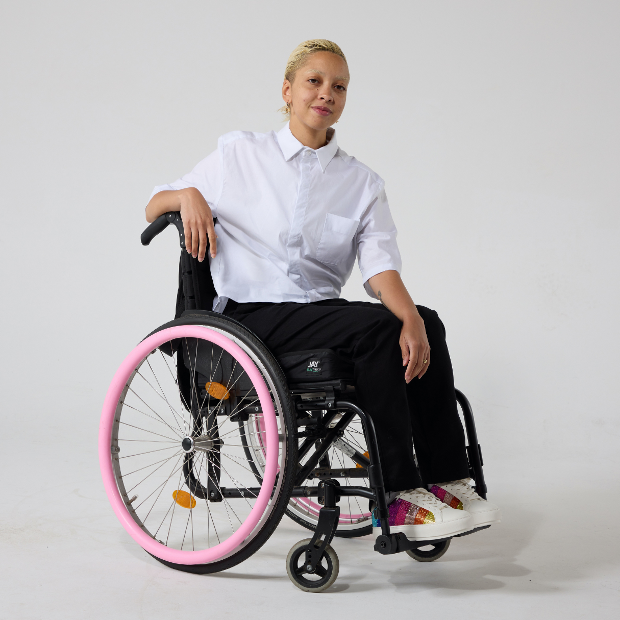A non-binary model of mixed heritage wears a white short sleeve hi-lo cotton shirt paired with black trousers and shoes with a rainbow patch on them. She is seated in a manual wheelchair with one of her hands resting on the back of the wheelchair while the other hand resting in her lap.