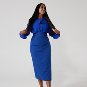 A South Asian woman with long glossy black hair is standing against an offwhite background. She is holding her hair slightly out either side, she is wearing a royal blue chiffon pussy bow tie shirt with a matching colour wrap skirt.