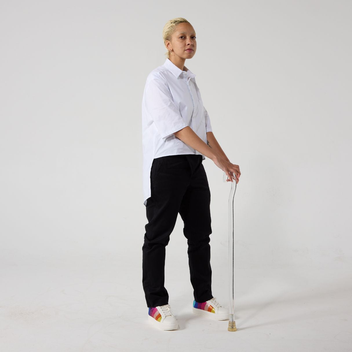 A non-binary model of mixed heritage is standing with a walking stick and looking at the camera, sideways. She is wearing black wrap twill trousers paired with a white shirt and sneakers. Her hands are resting on handle of the clear walking stick.