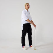 A non-binary model of mixed heritage is standing with a walking stick and looking at the camera, sideways. She is wearing black wrap twill trousers paired with a white shirt and sneakers. Her hands are resting on handle of the clear walking stick.