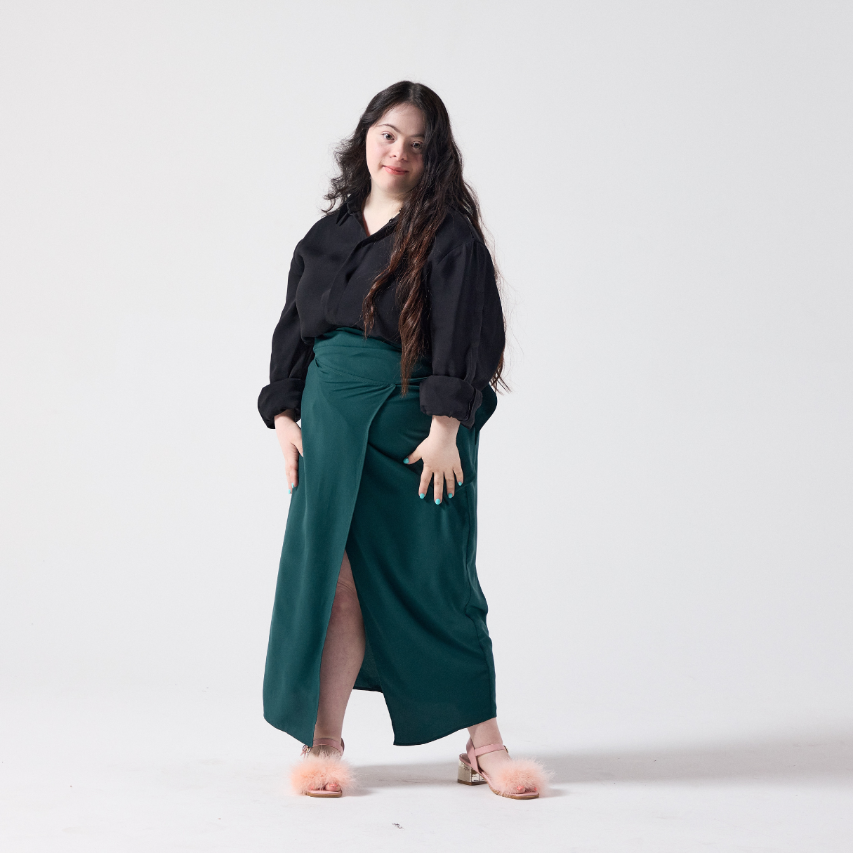 A white model with long dark brown hair is smiling at the camera. She is wearing a wrap skit in forest green paired with a black shirt and her own heels. She is standing with her legs posed apart and her hands resting on her sides.