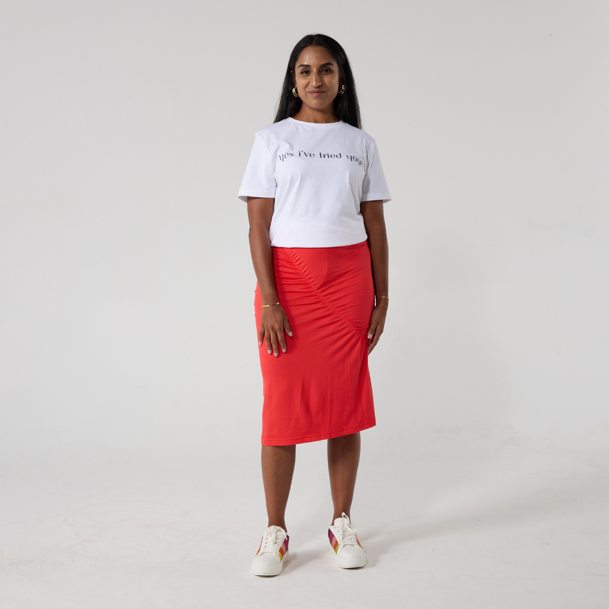 A south asian woman with long black hair is smiling at the camera. She is wearing a  coral red ruched skirt with a white t shirt and white sneakers. She is casually standing with her hands close to her side.