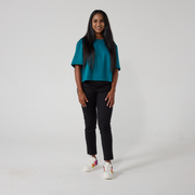 A south asian model with long black glossy hair is smiling at the camera. She is wearing black wrap twill trousers with a teal top and white sneakers. She is standing with her hands relaxed on her sides.