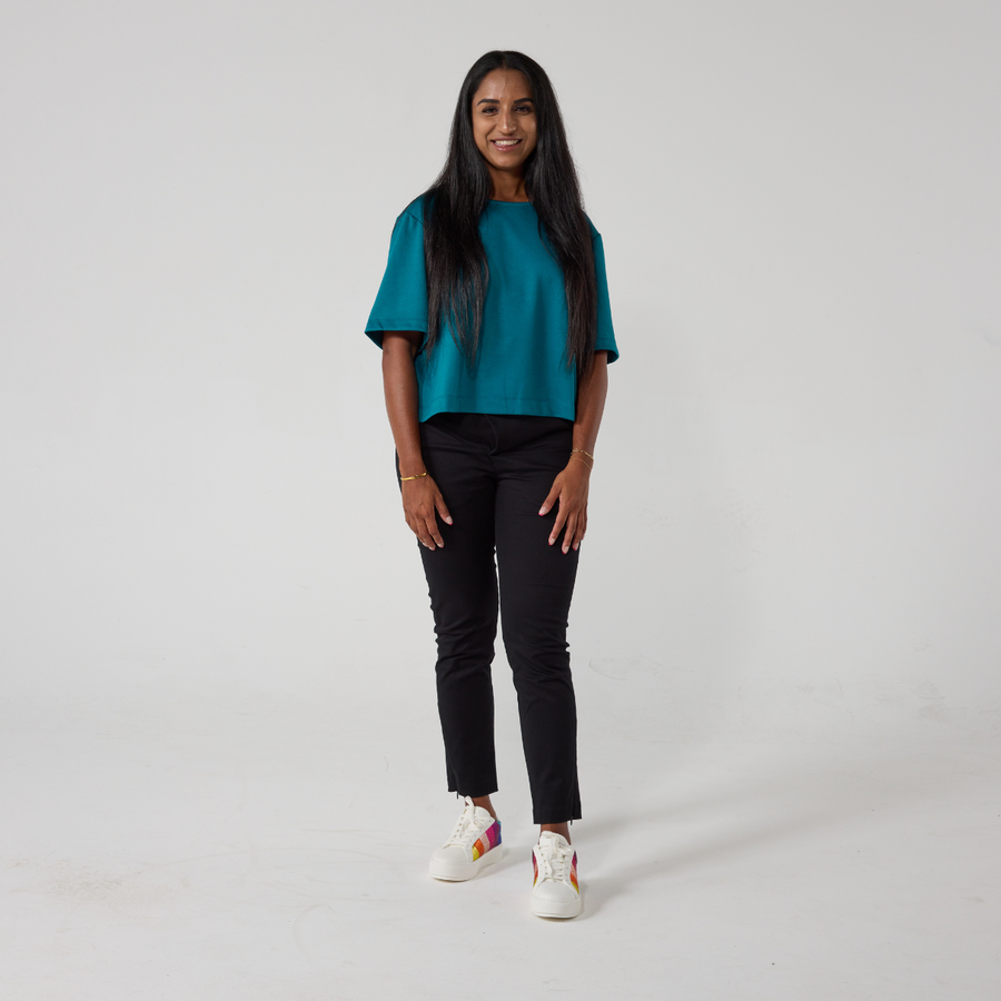 A south asian model with long black glossy hair is smiling at the camera. She is wearing black wrap twill trousers with a teal top and white sneakers. She is standing with her hands relaxed on her sides.