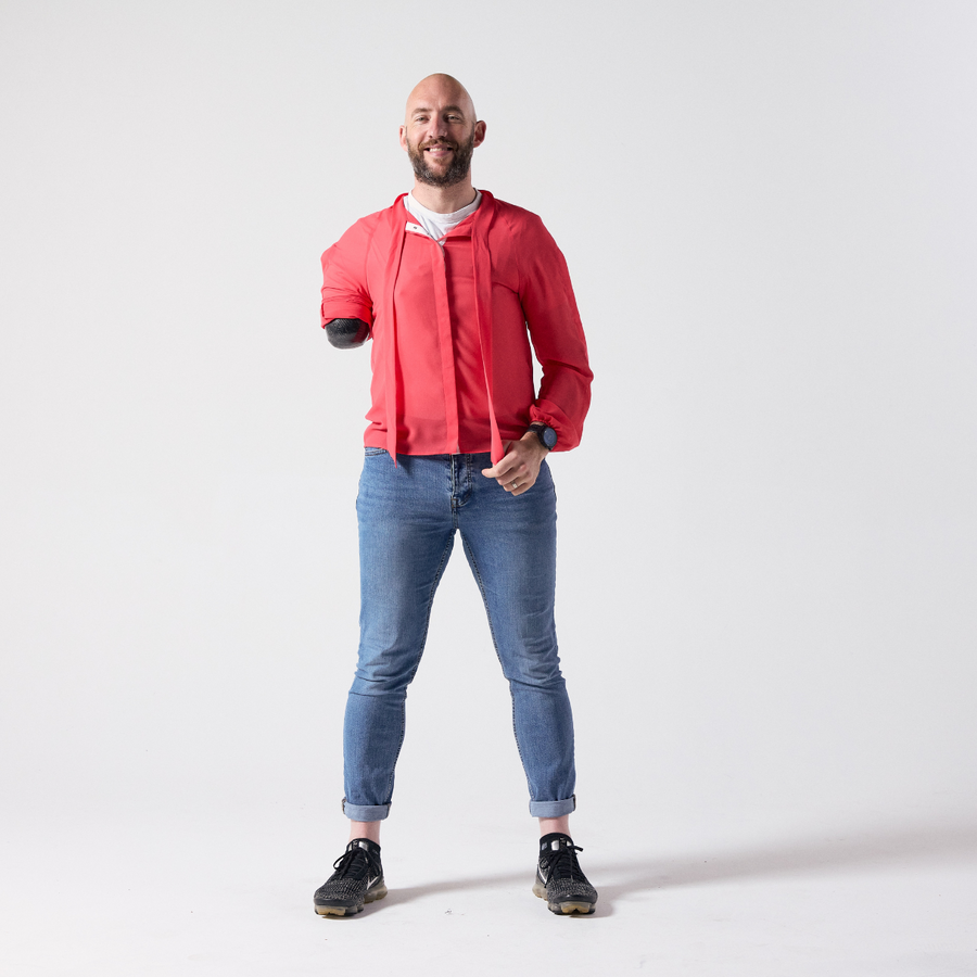 A smiling white man with a brown grey beard is standing while facing the camera. He is wearing a red pussy bow shirt with an untied knot paired with his own jeans and shoes.