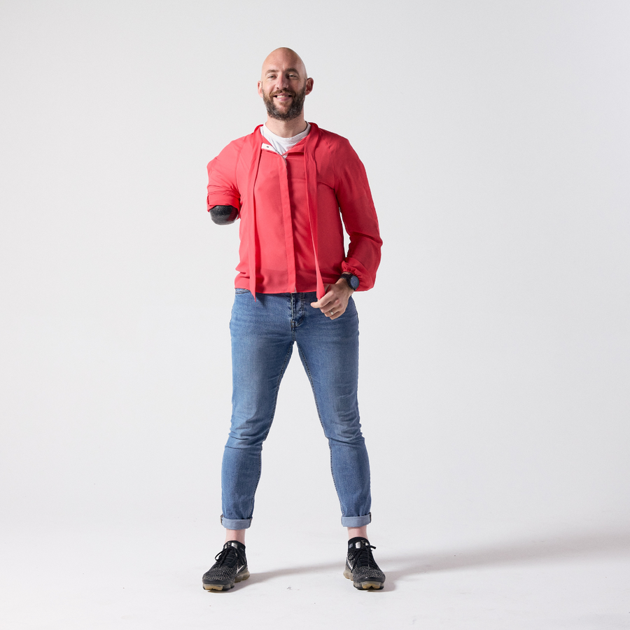 A smiling white man with a brown grey beard is standing while facing the camera. He is wearing a red pussy bow shirt with an untied knot paired with his own jeans and shoes. 