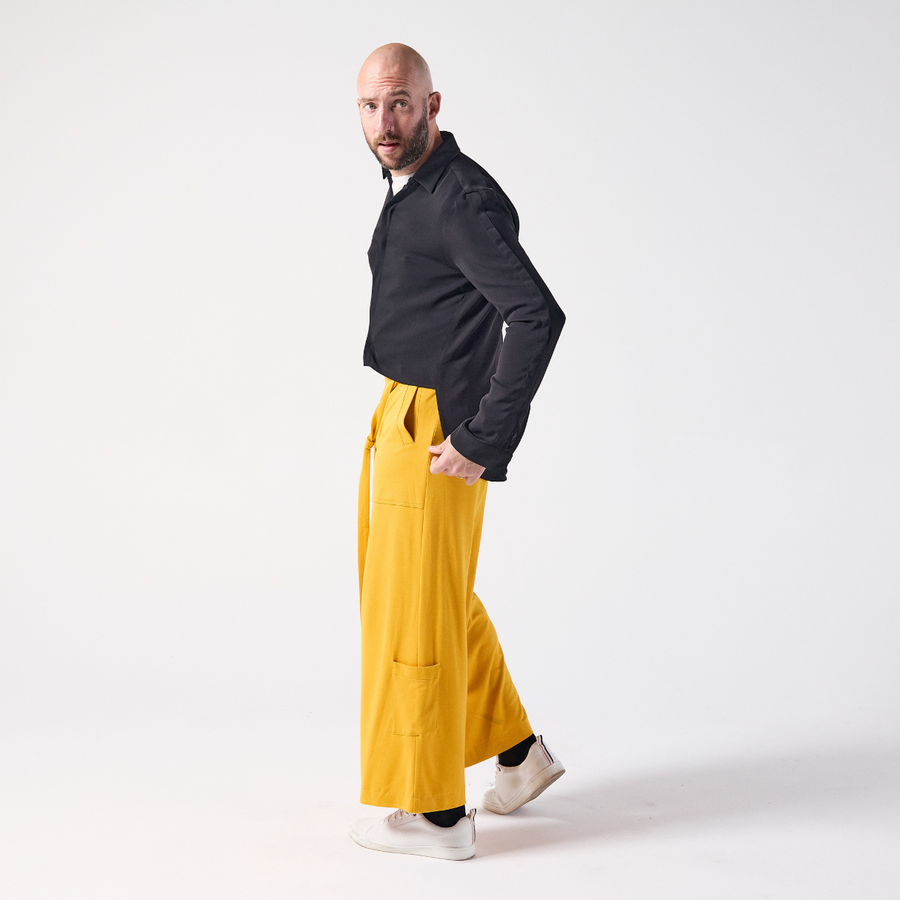 A white man is posing while moodily looking away from the camera. He is wearing jersey culottes in mustard yellow paired with a black shirt and white sneakers. He is standing while holding the edge of his shirt in one hand.