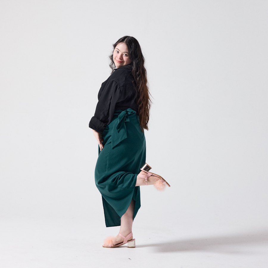 A white woman with long dark brown hair is smiling at the camera. She is wearing a forest green wrap skirt with a black shirt and heels. She is posing with one leg up and hands close to her thighs.