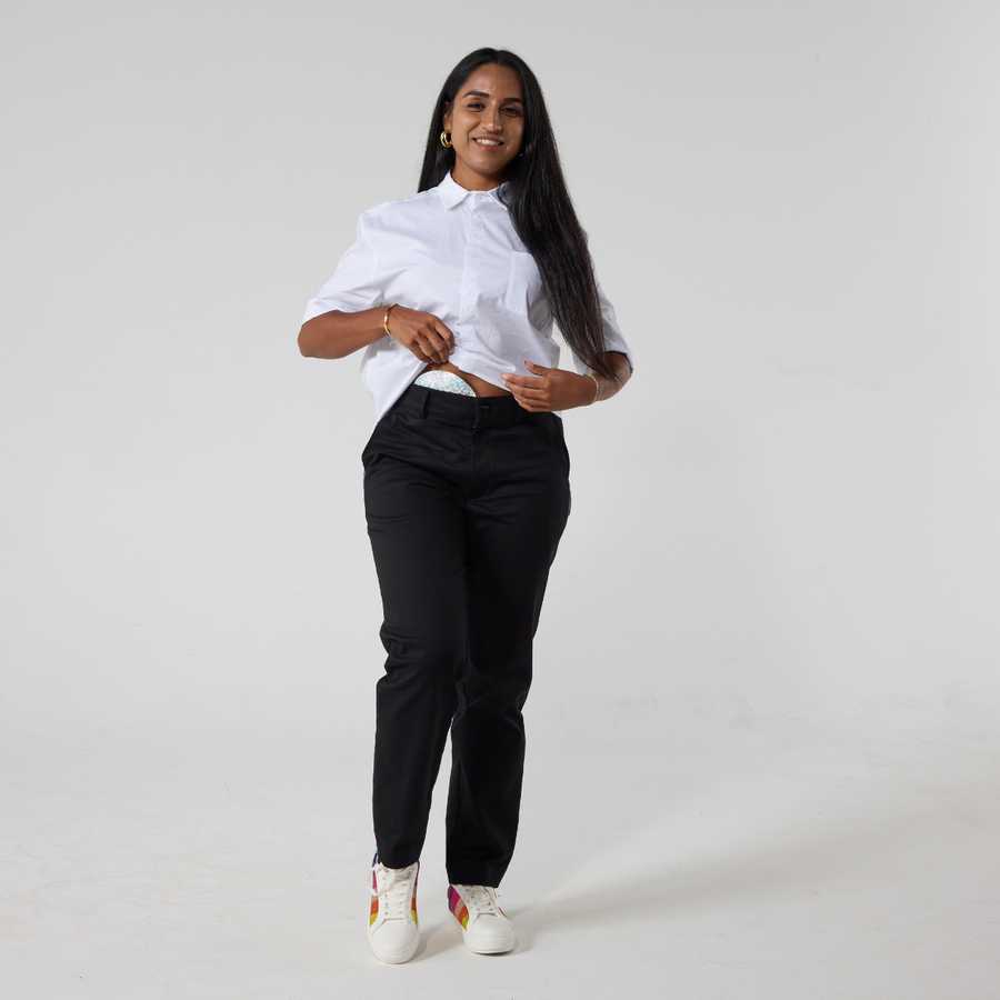 A South asian woman with long back hair is wearing black twill trousers in regular fit paired with a white shirt and sneakers with rainbow patches. She is lifting up her shirt hem to show her stoma bag and the accessibility of the trousers.
