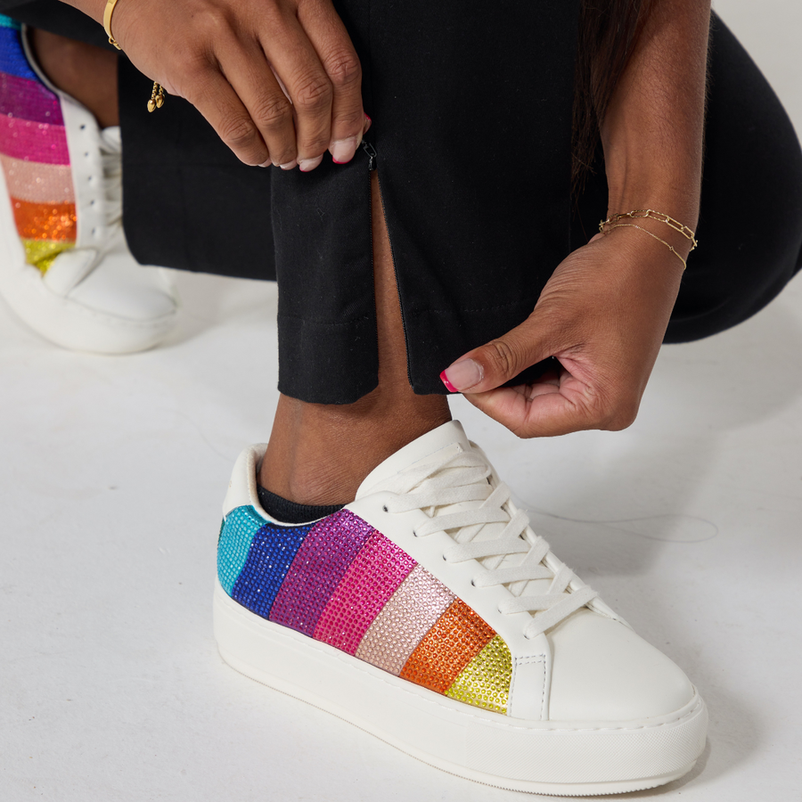 Closeup of a south asian model unzipping the side seam zipper at the hem of black twill trousers. She is wearing white sneakers with rainbow patches on either side.