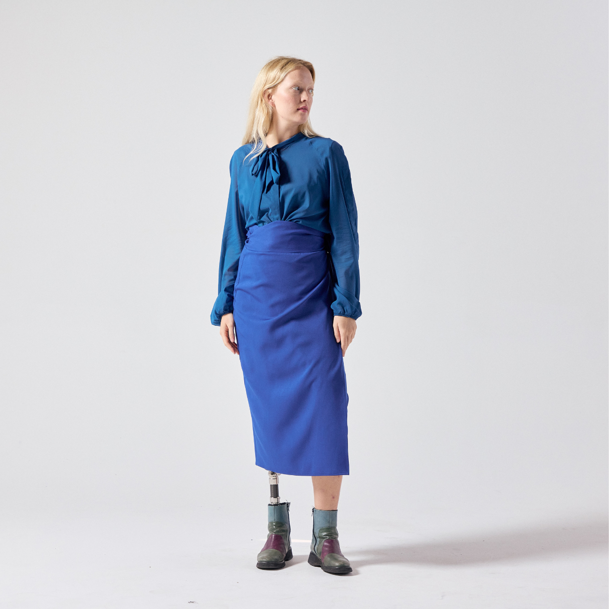 A white woman with long blonde hair is posing while looking away from the camera. She is wearing cobalt blue wrap skirt paired with a teal shirt and her own boots. She is standing with her hands relaxed to her sides.