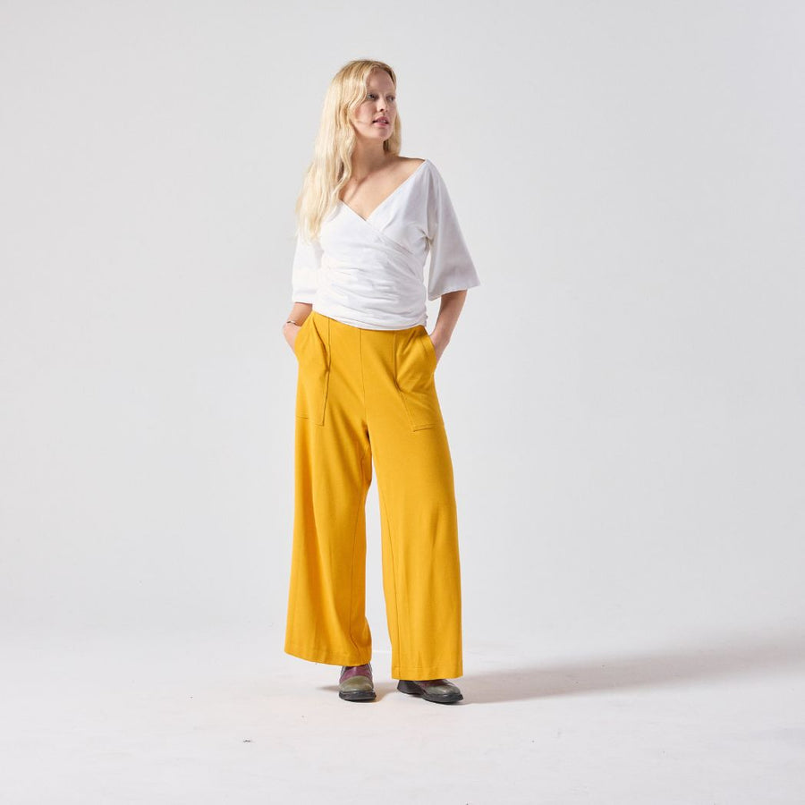 A white woman with blonde hair poses standing, looking away from the camera. She is wearing a white Jersey wrap top paired with yellow culottes and her own boots. She is standing with her hands in her pockets against a light grey background.