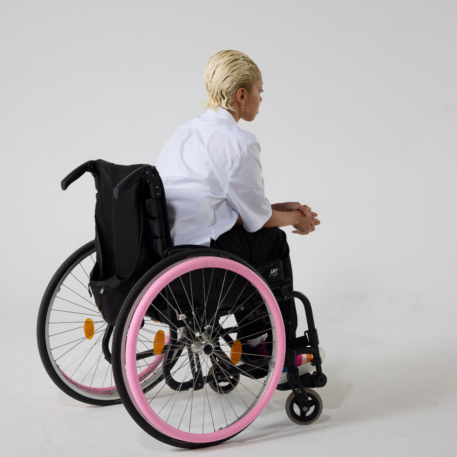 A non-binary model of mixed heritage wears a white short sleeve hi-lo cotton shirt paired with black trousers and shoes with a rainbow patch on them. She is seated in a manual wheelchair with her back facing the camera. She has gelled down blonde hair and her hands are resting on her knees.