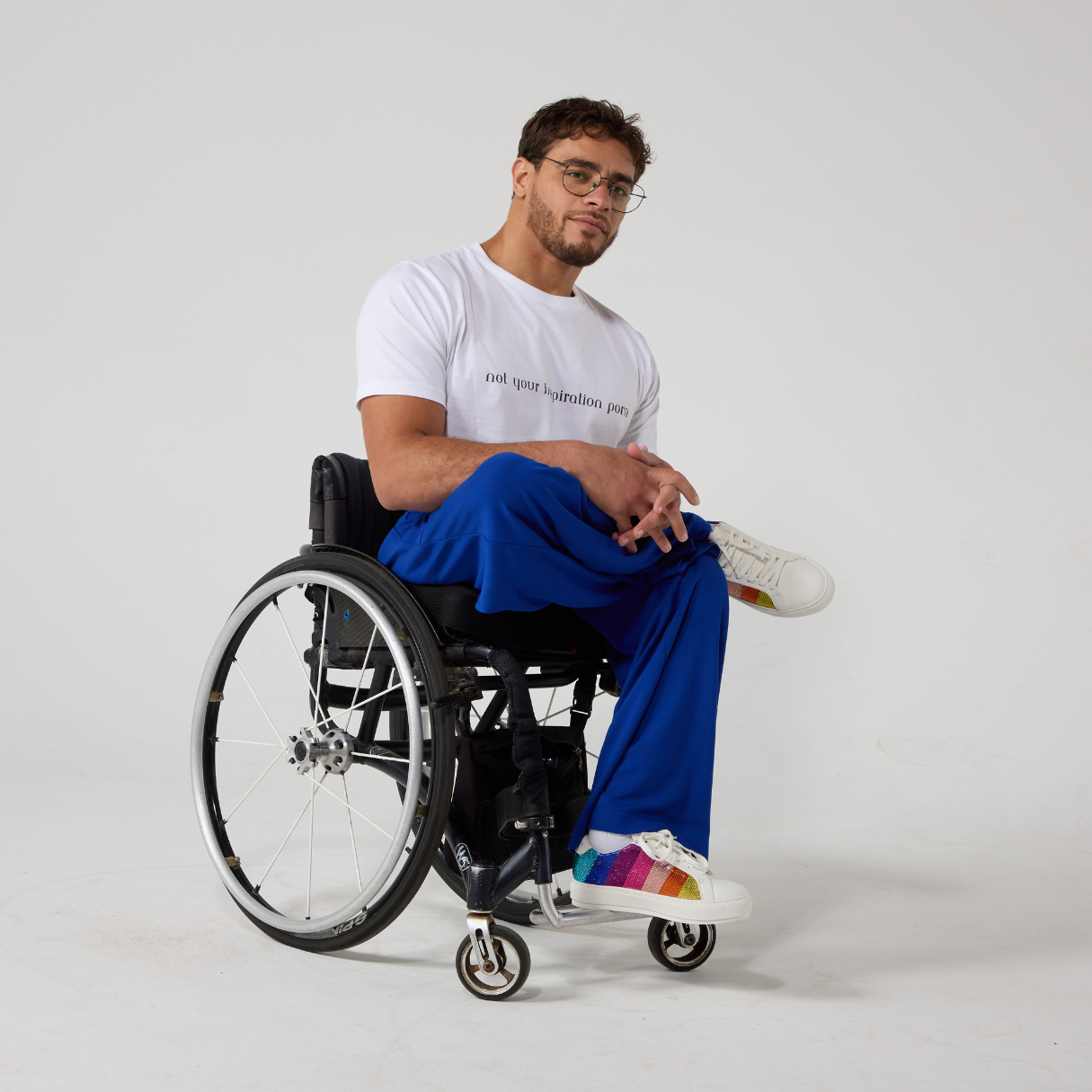 A male model of mixed heritage is looking at the camera. He is wearing royal blue culottes with a white t-shirt and white sneakers. He is sitting in a manual wheelchair with his leg crossed and his hands resting in his laps.