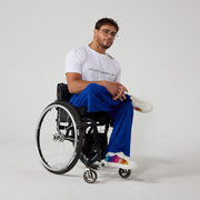 A male model of mixed heritage is looking at the camera. He is wearing royal blue culottes with a white t-shirt and white sneakers. He is sitting in a manual wheelchair with his leg crossed and his hands resting in his laps.