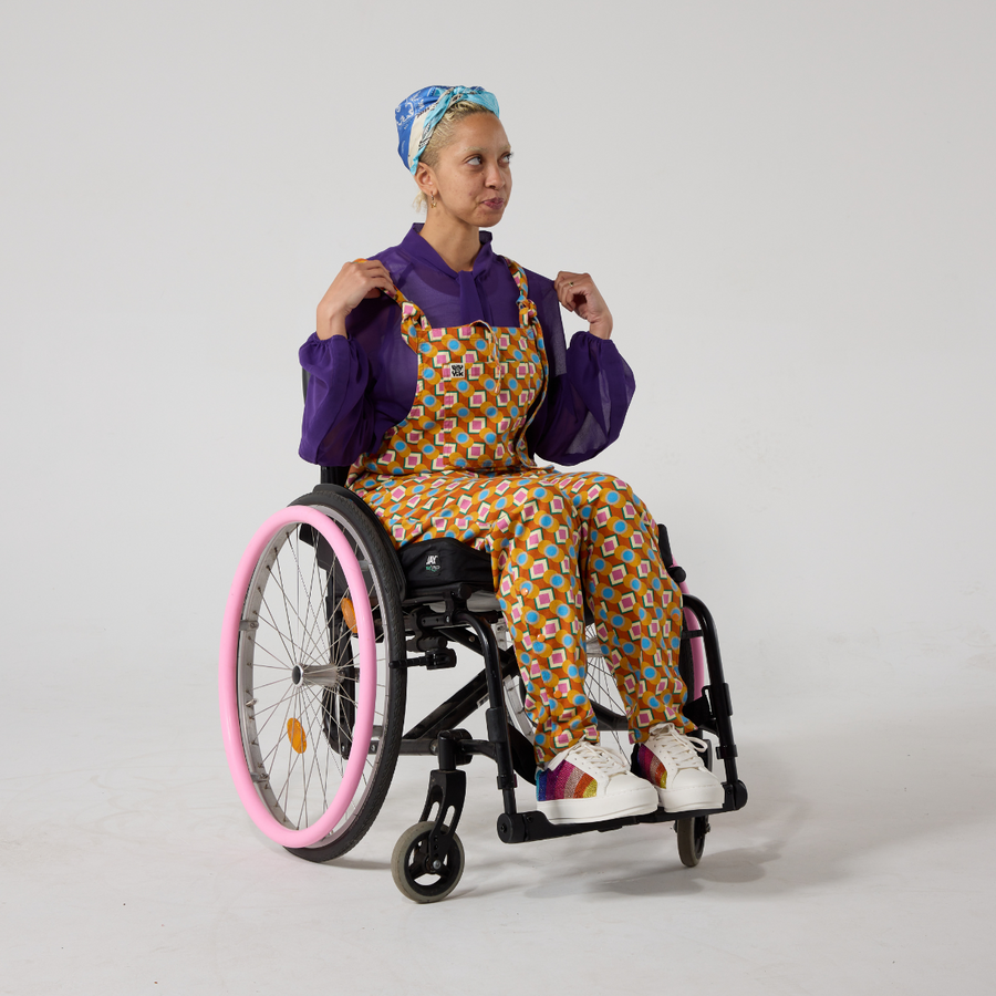 A non binary model of mixed heritage wears a purple pussy bow tie shirt under brightly orange pattern dungarees. They are a manual wheelchair user with dyed blonde hair and a blue silk headscarf. They have rainbow accented sneakers on.
