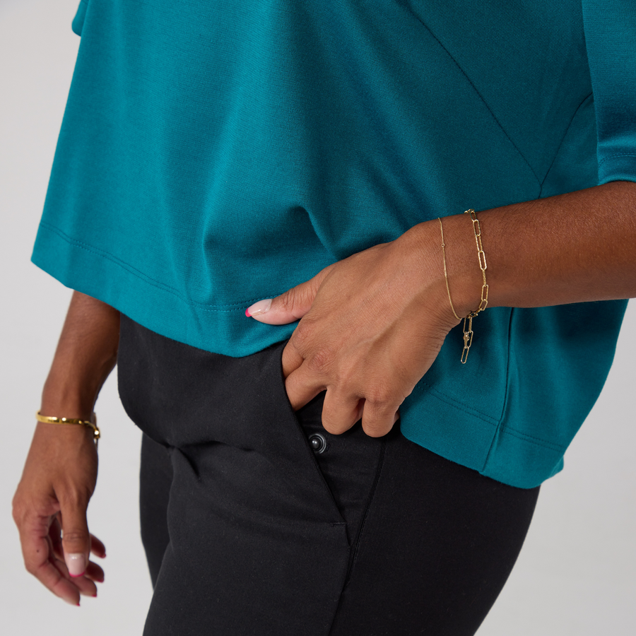 Close-up of the backless teal top highlighting the hemline. It is modelled on a south asian woman. One of her hands is in her pocket while the other is relaxed to her side.