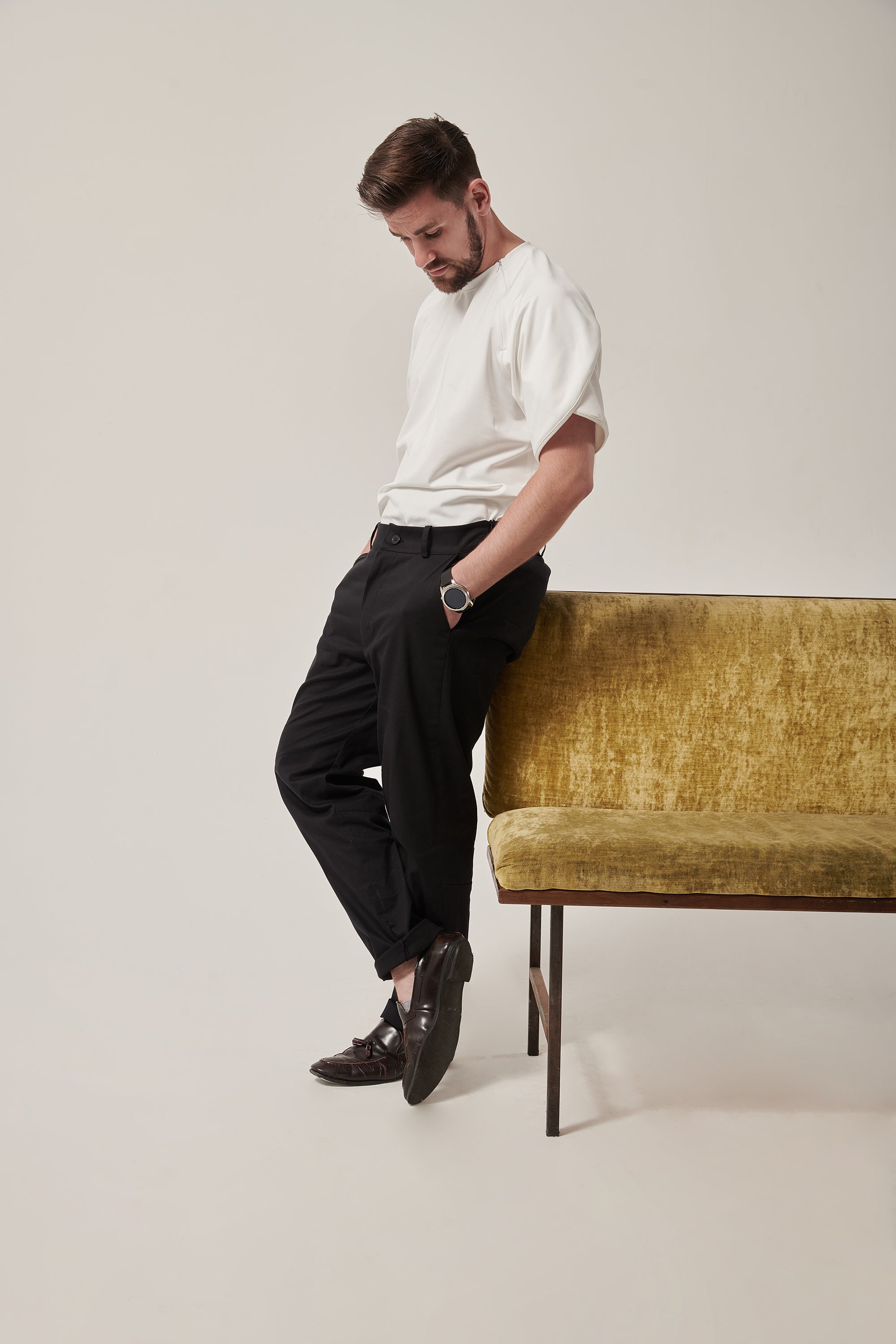 A white man stands leaning against a crushed velvet moss coloured sofa, his legs crossed and arms in his pockets. He is wearing a white t-shirt with a wrap sleeve. There are concealed zips to allow chest access at the front.