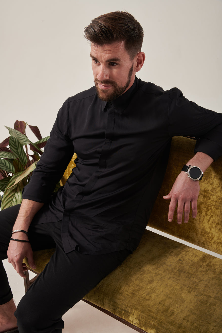 A white man with styled dark brown hair looks off camera. He reclines on a crushed moss coloured velvet sofa. There is a plant at his right side. The background is white.