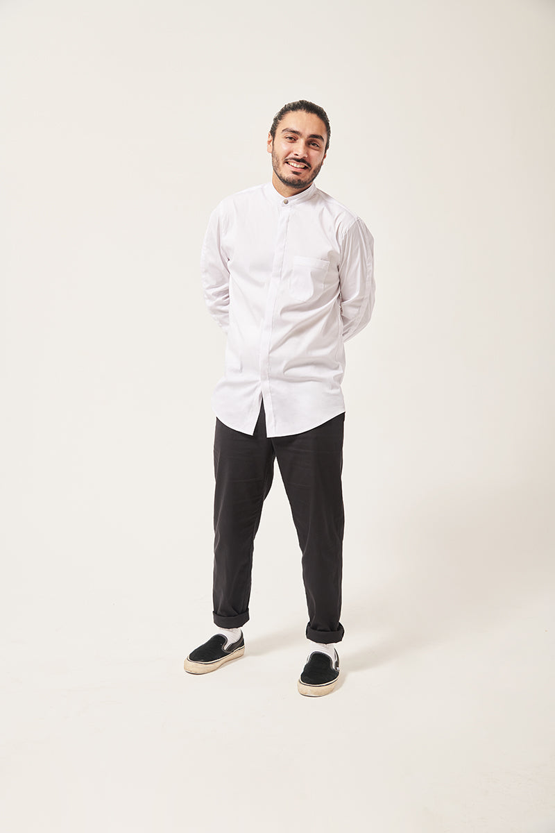 A south Asian man laughs towards he camera. He is wearing a white shirt with a mandarin collar and a pocket on the left chest. HIs arms are behind his back, his legs are apart. He is wearing black vans. The background is shades of grey.