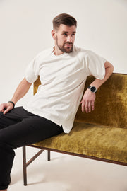 A white man with styled dark brown hair looks to the right. He is reclining on a crushed velvet moss coloured sofa. He is wearing a white t-shirt with wrap sleeves and concealed front zips to allow chest access. His left arm is resting on the back of the sofa and he has a large black watch on.