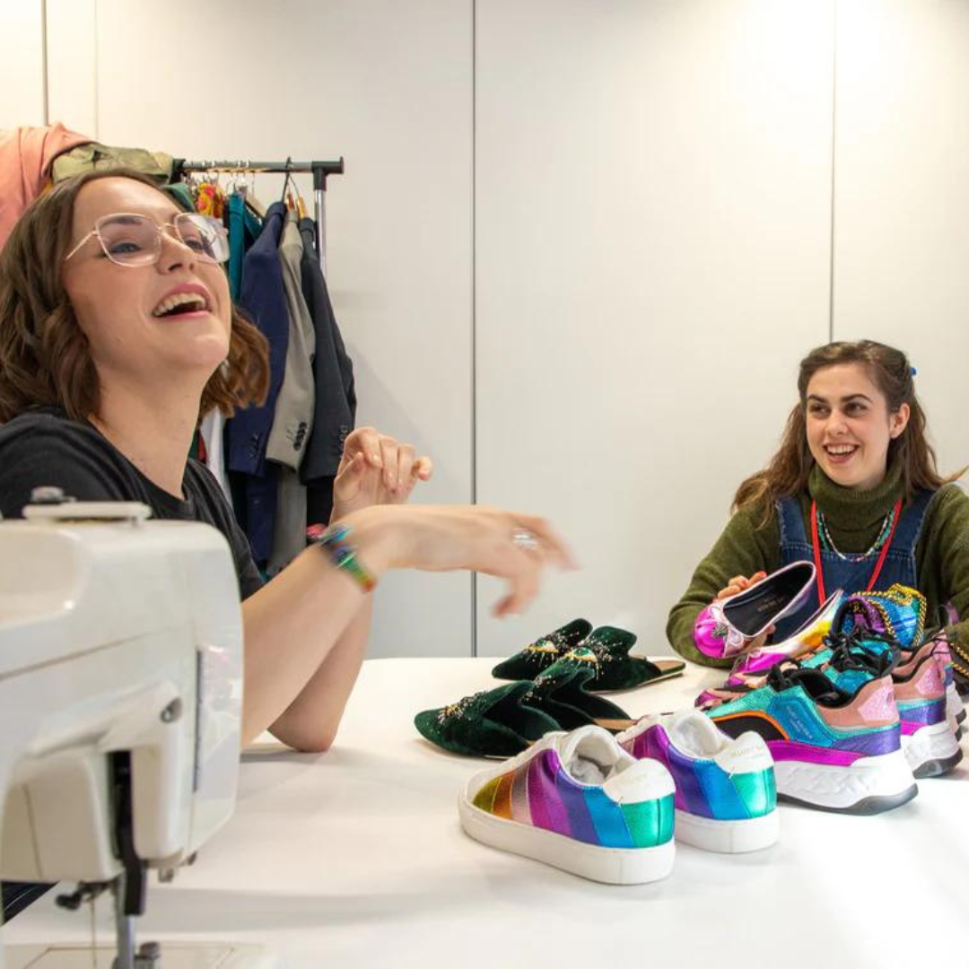Two women are sitting at a desk. There is a sewing machine and an array of rainbow coloured shoes and bags on the table. The women sitting next to the sewing machine is wearing glasses and tilting her head back laughing
