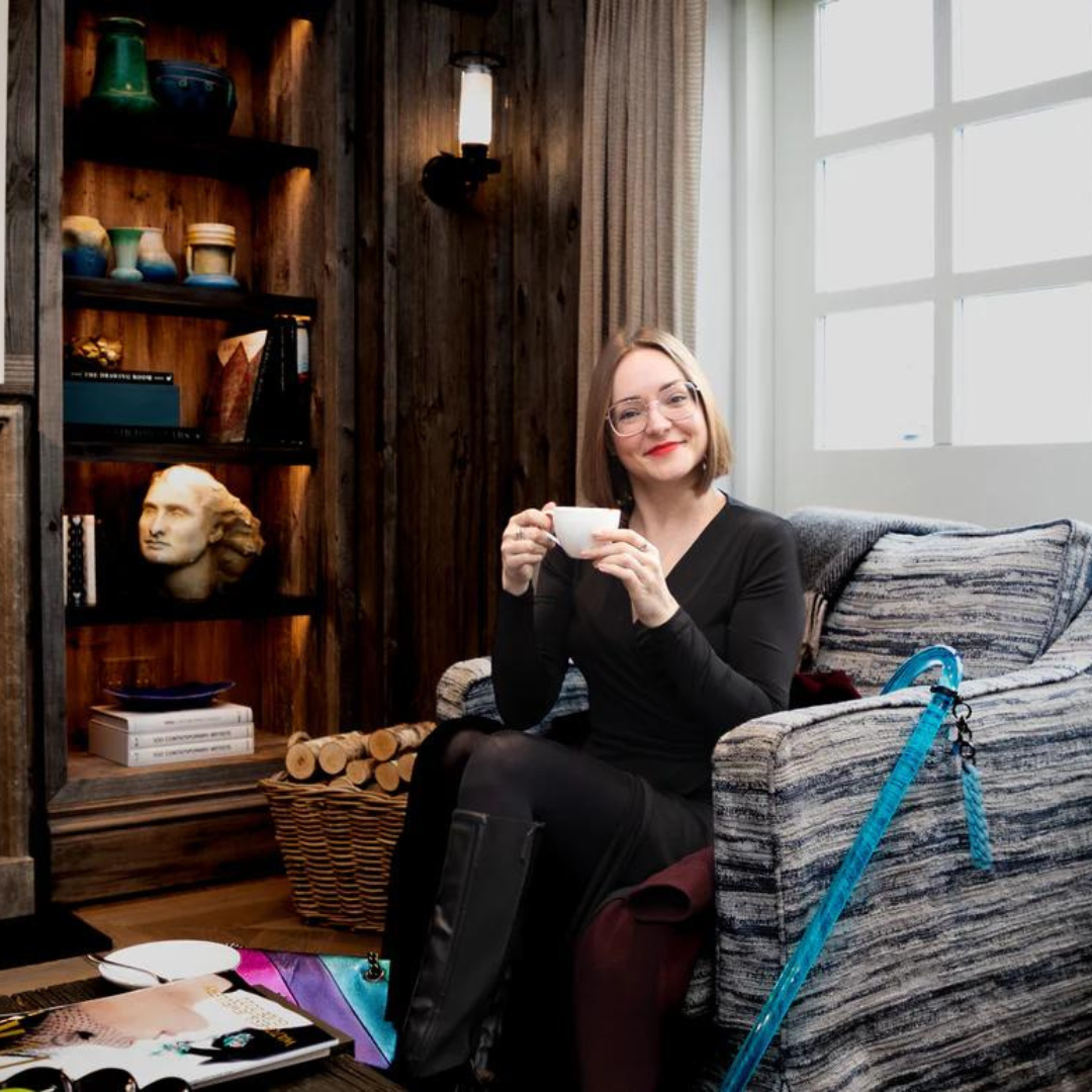 Victoria, white woman with brown hair, is sitting in a brown chair with her blue mobility aide balanced on the arm. She wearing a black jumper and trousers, is holding a cup of tea and smiling to the camera.