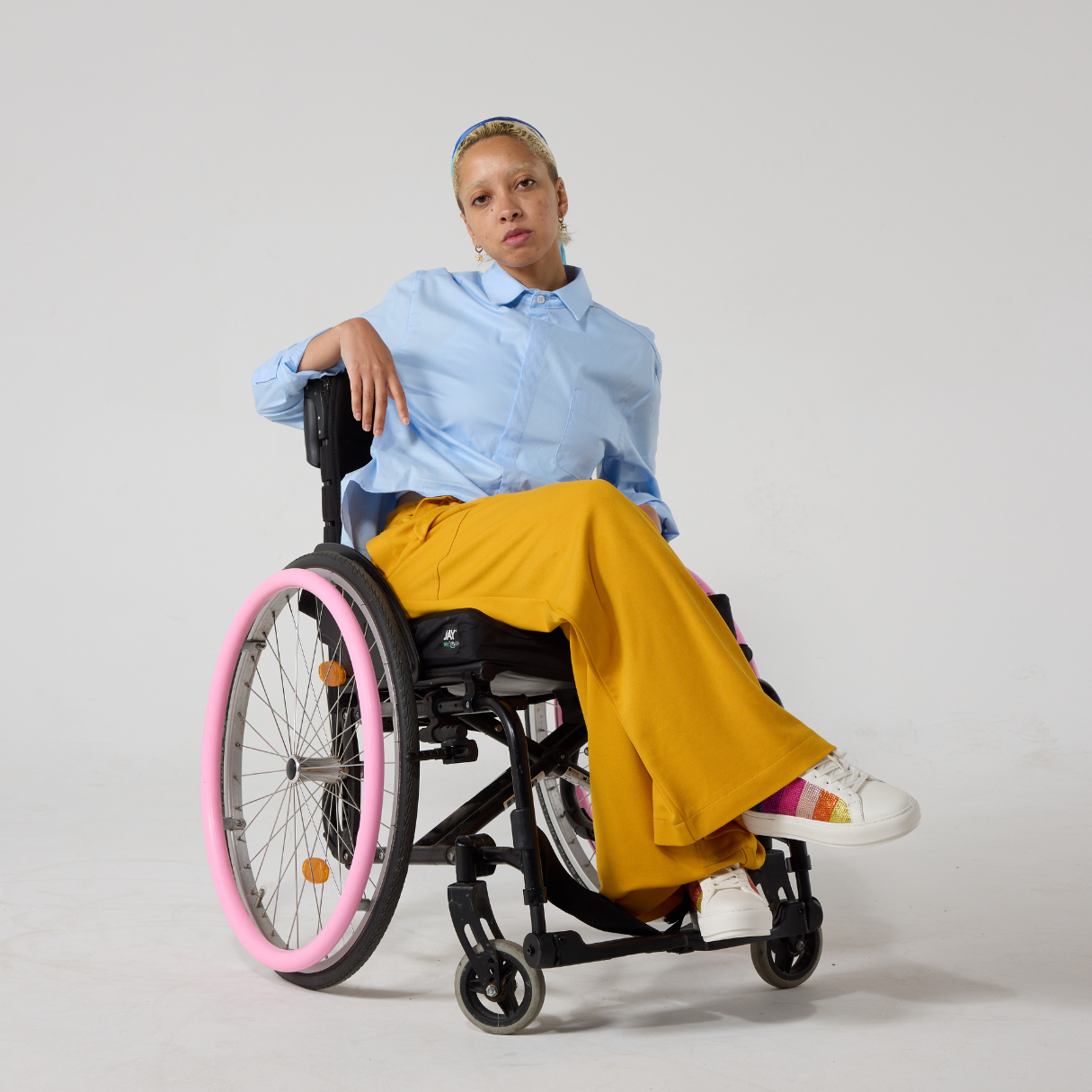 Junior is mixed heritage and has dyed blonde afro hair under a blue silk scarf. They are seated in a wheelchair, wearing a light blue shirt with a collar that has a pocket on the left side as worn. It is cropped at the front. They also have mustard colour jersey culottes on with white sneakers that have sparkly rainbow accents. Set against an off white back ground.