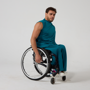 Model is male presenting of mixed heritage. They are wearing a sleeveless teal coloured jersey top that has a high neck, with matching teal jersey culottes. They are a manual wheelchair user, against an off white back ground.