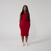 Tanya is a South Asian woman with long black glossy hair. She is stood against an off white back ground wearing a raspberry red dress that has an over lay on the top half and a keyhole opening. She is standing with her hands in her pockets, wearing white sneakers.
