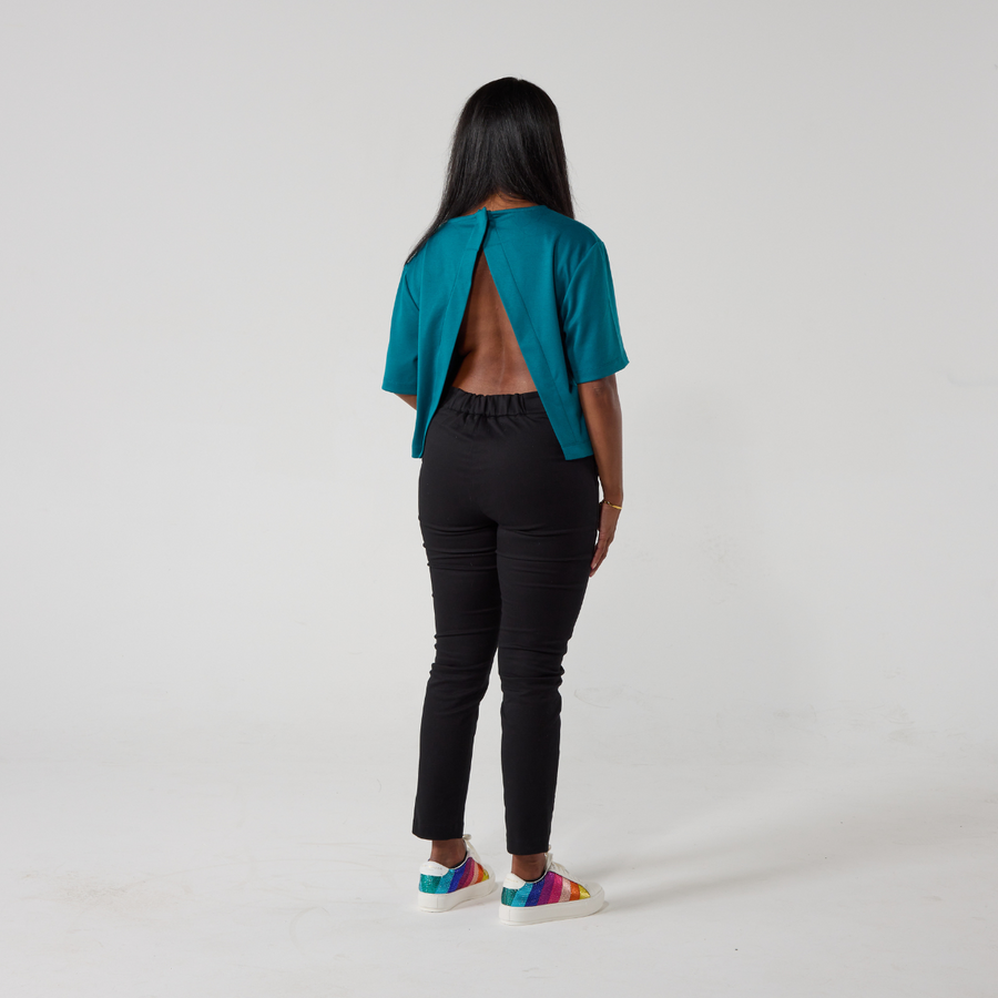 A south asian model with long black glossy hair is standing with her back facing the camera. She is wearing black wrap twill trousers with a teal top and white sneakers. She is standing with her hands relaxed on her sides.