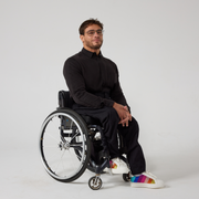 A male model of mixed heritage is looking at the camera. He is wearing black seated twill trousers in regular fit paired with a black shirt and white sneakers. He is sat in a manual wheelchair with his hands resting in his lap.