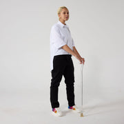 A non-binary model of mixed heritage is standing with a walking stick and looking at the camera, sideways. She is wearing black wrap twill trousers paired with a white shirt and sneakers. Her hands are resting on handle of the clear walking stick. 