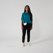 Model is female presenting and is south asian. They are wearing a teal coloured cropped t-shirt style top, black trousers and the lower half of a sparkly stoma cover is also visible. They are against an off white background.