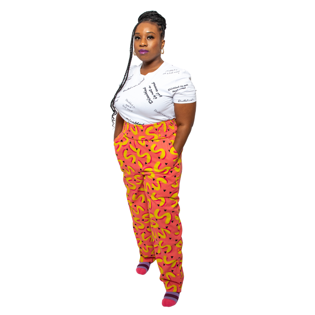 Glynis, a black woman with an arm limb difference, is wearing pink and yellow vibrant , high waisted trousers with a white tshirt with the text slogans "Disabled is not a bad word" .