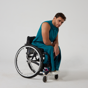 Model is male presenting of mixed heritage, leaning forward and resting their arms across their knees. They are wearing a sleeveless teal coloured jersey top that has a high neck, with matching teal jersey culottes. They are a manual wheelchair user, against an off white back ground.