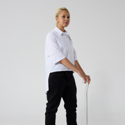 A non-binary model of mixed heritage wears a white short sleeve hi-lo cotton shirt paired with black trousers. She's standing with the help of a clear walking stick with her hands resting on the handle of the stick.