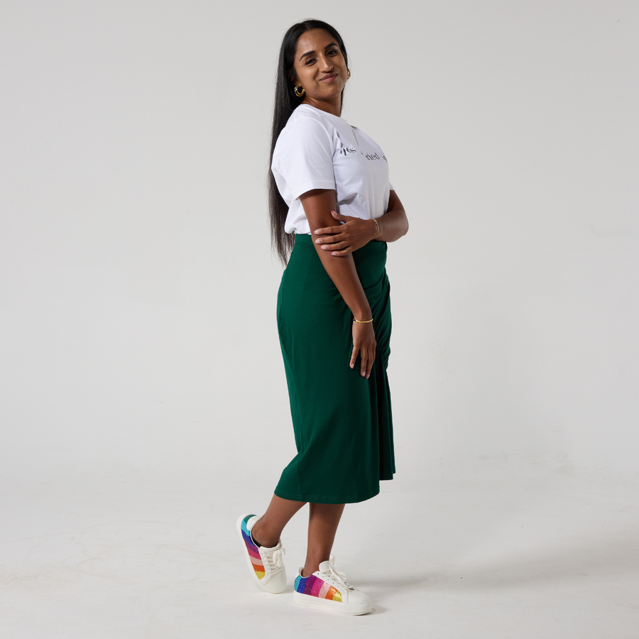 Side view of a south asian model with long black hair smiling at the camera. She is wearing a forest green ruched skirt paired with a white t shirt and white sneakers. She is standing with her feet crossed.