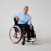 A  non-binary model of mixed heritage is looking at the camera. She is wearing seated black twill trousers with a blue shirt and white sneakers. She is sat in a manual wheelchair with one of her hands resting on the chair's back and the other resting on her lap.