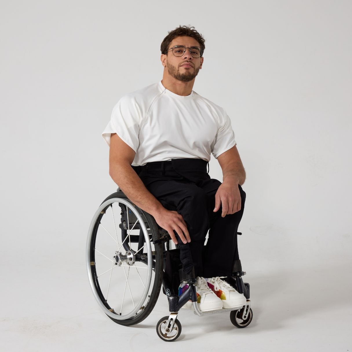 A male model of mixed heritage is looking at the camera. He is wearing black seated 'wrap' twill trousers in regular fit paired with a white t-shirt and white sneakers. He is sat in a manual wheelchair with one of his hands on his knee while the other by his side.