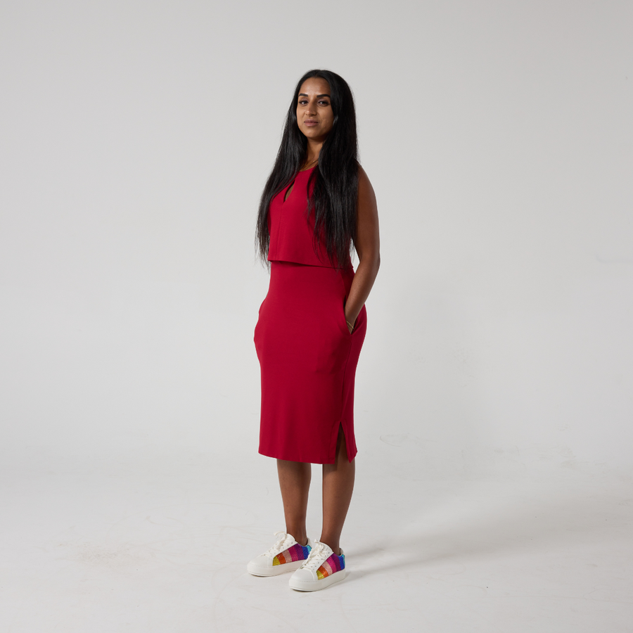 Tanya is a South Asian woman with long black glossy hair. She is stood against an off white back ground wearing a raspberry red dress that has an over lay on the top half and a keyhole opening. She is standing with her hands in her pockets, wearing white sneakers. She is angled slightly to the side so the rainbow sparkles can be seen on her shoes.