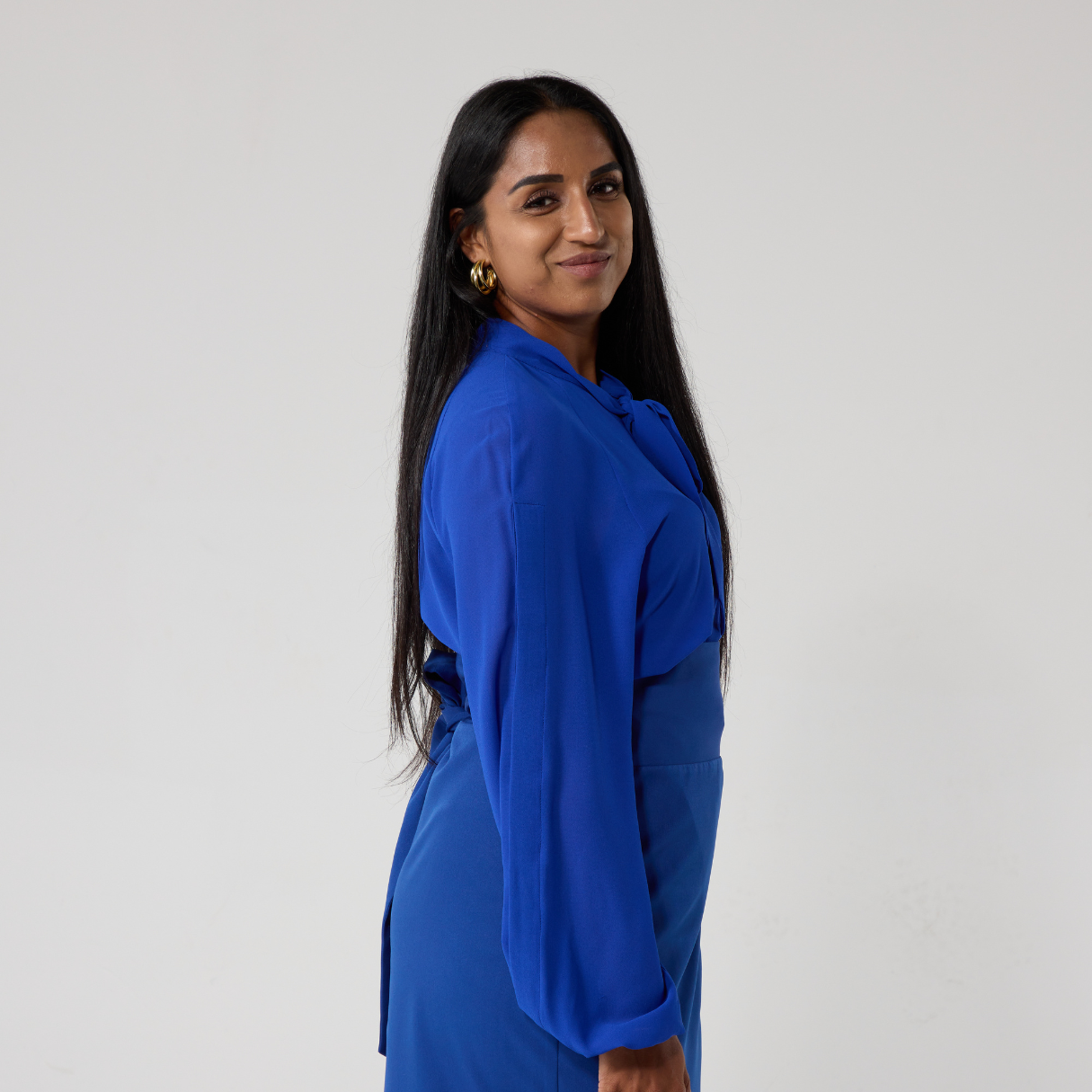 A south asian woman shows a side profile of her body whilst turning her head to look at the camera. She is wearing a royal blue chiffon shirt, there is a concealed opening down the length of the sleeve which has a slight bell shape to its hem.