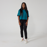 A south asian model with long black glossy hair is smiling at the camera. She is wearing black wrap twill trousers with a teal top and white sneakers. She is standing with her hands relaxed on her sides. 
