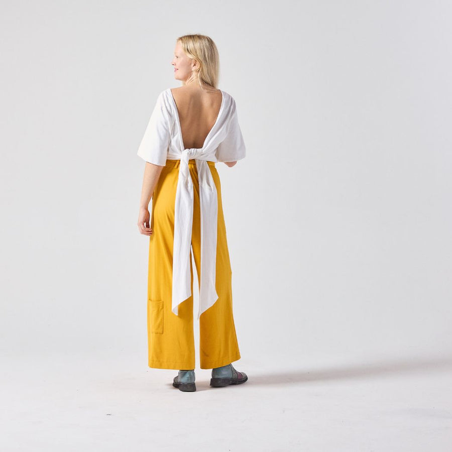 A blonde white woman standing with her back facing the camera while looking sideways. She is wearing a short sleeve white wrap top paired with yellow culottes and her own boots. The back panels of the wrap top are tied at her waist.