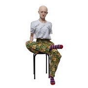 Hannah, a white woman, is wearing green and yellow high waisted trousers in a floral print with a white tshirt with black text reading "Disabled is not a bad word". She is sitting on a black stool with one foot across her knee