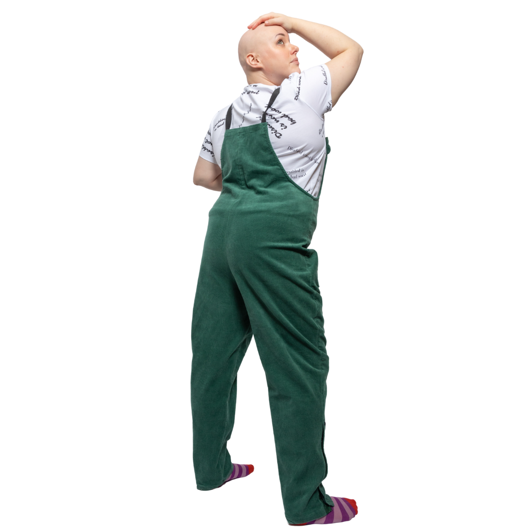 Laura, a white woman with alopecia, is standing facing away from the camera with her hand on her head. She is wearing green dungarees with a white t shirt underneath reading "disabled is not a bad word" with stripy purple and pink socks