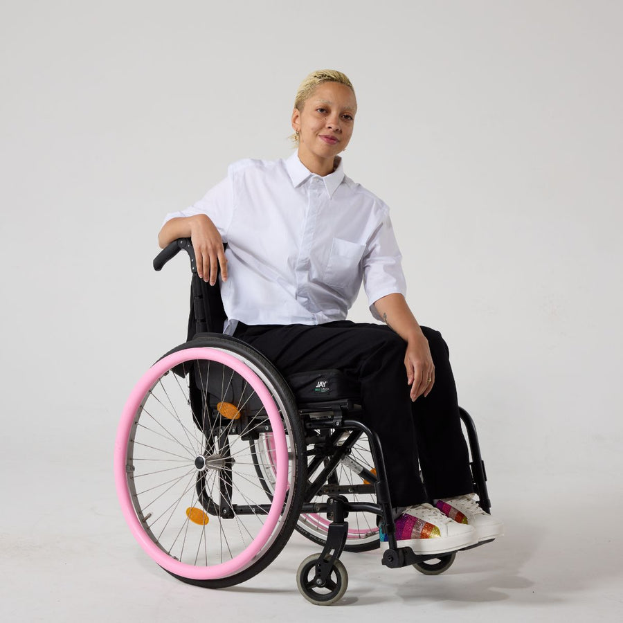 A non-binary model of mixed heritage is smiling at the camera. She is wearing seated black wrap twill trousers with a white shirt and white sneakers. She is sat in a manual wheelchair with one of her hands resting on the chair's back and the other resting on her lap.